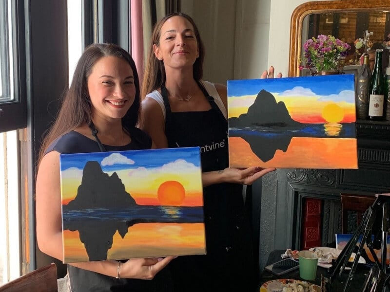 Two women stand by a window holding finished sunset landscape paintings